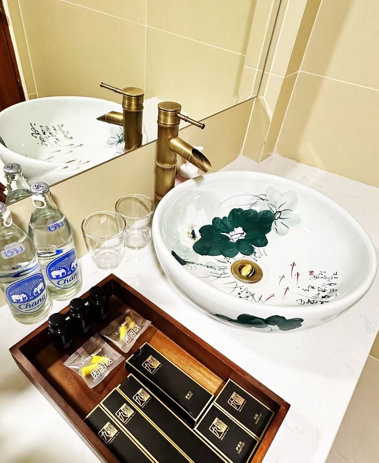 A bathroom with a white sink, two faucets, and a mirror. There are bottles, glasses, and a tray with toiletries.