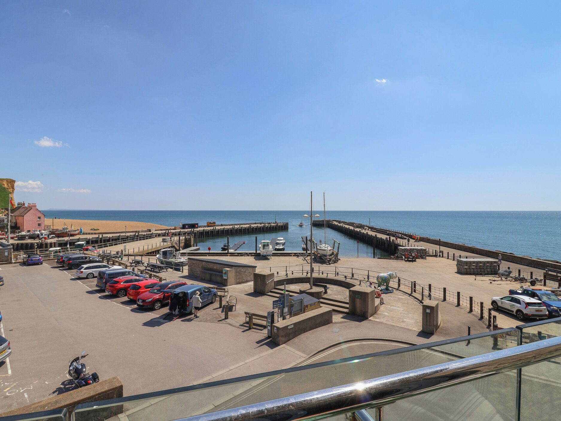 A coastal marina with boats docked, a parking area with cars, and a clear view of the sea.
