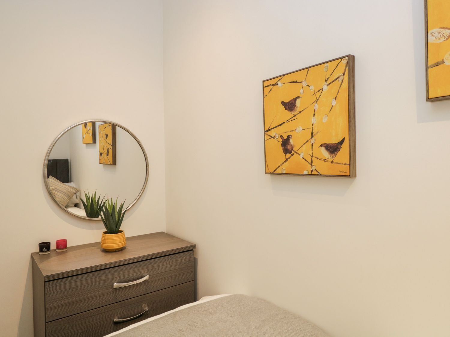 A bedroom with a round mirror, a bedside table with a plant, and two framed artworks on the wall.