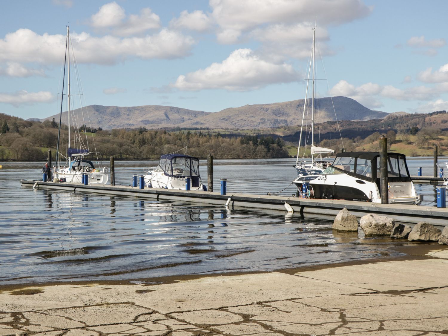 A calm lake with boats docked at a pier, surrounded by mountains and trees.