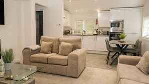 Una sala de estar moderna con un sofá color beige, una mesa de centro de vidrio y un comedor con una mesa y sillas. Se ve la cocina al fondo, con gabinetes blancos y electrodomésticos de acero inoxidable.