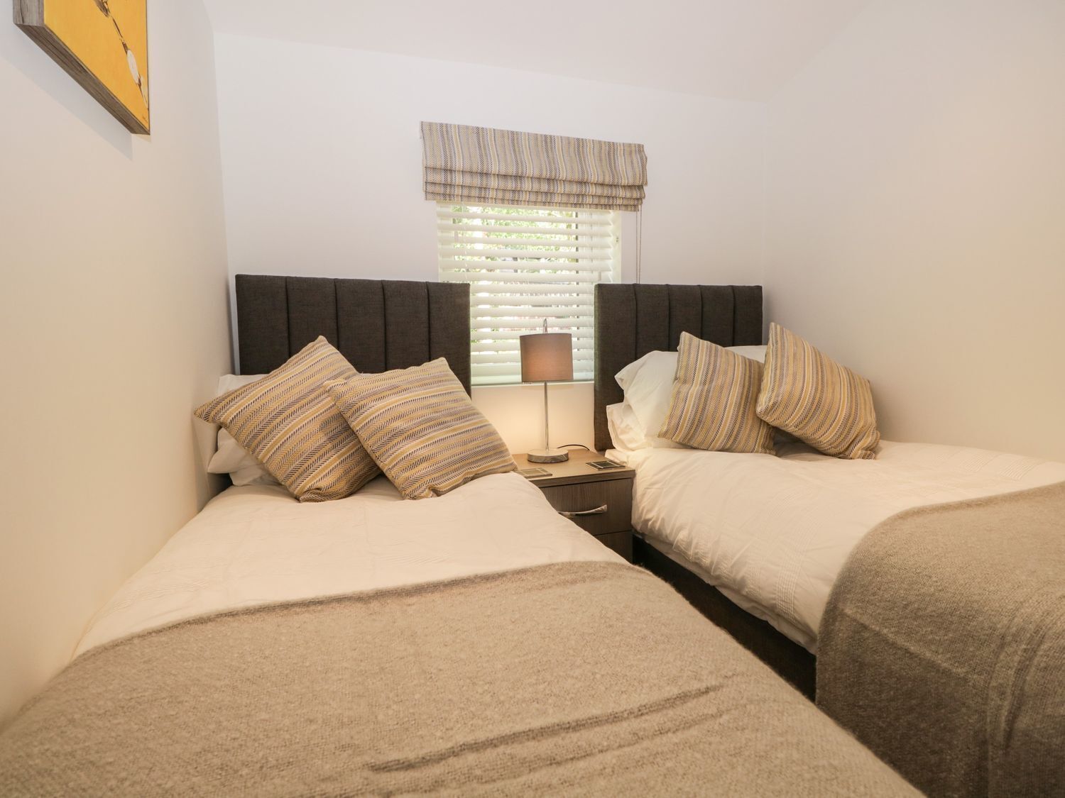 A twin room with two beds, a nightstand, a lamp, and a window with blinds.