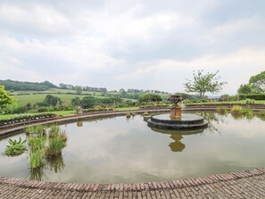 Ein runder Teich mit einer Fontäne in der Mitte, umgeben von einem Pflasterweg und üppigem Grün.