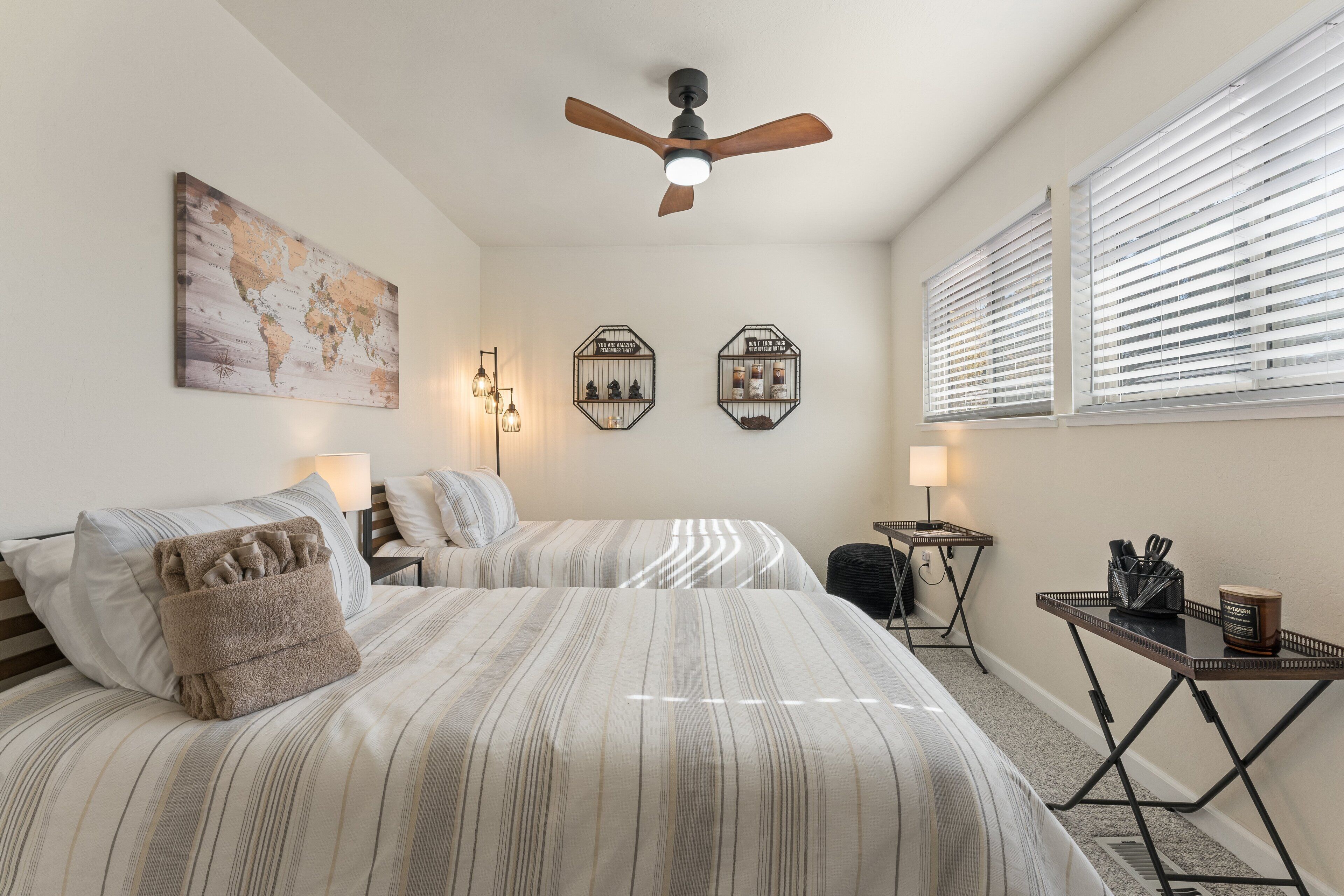 A hotel room with two beds, a ceiling fan, a world map, hexagonal wall decorations, and a small table with a lamp and a tray.