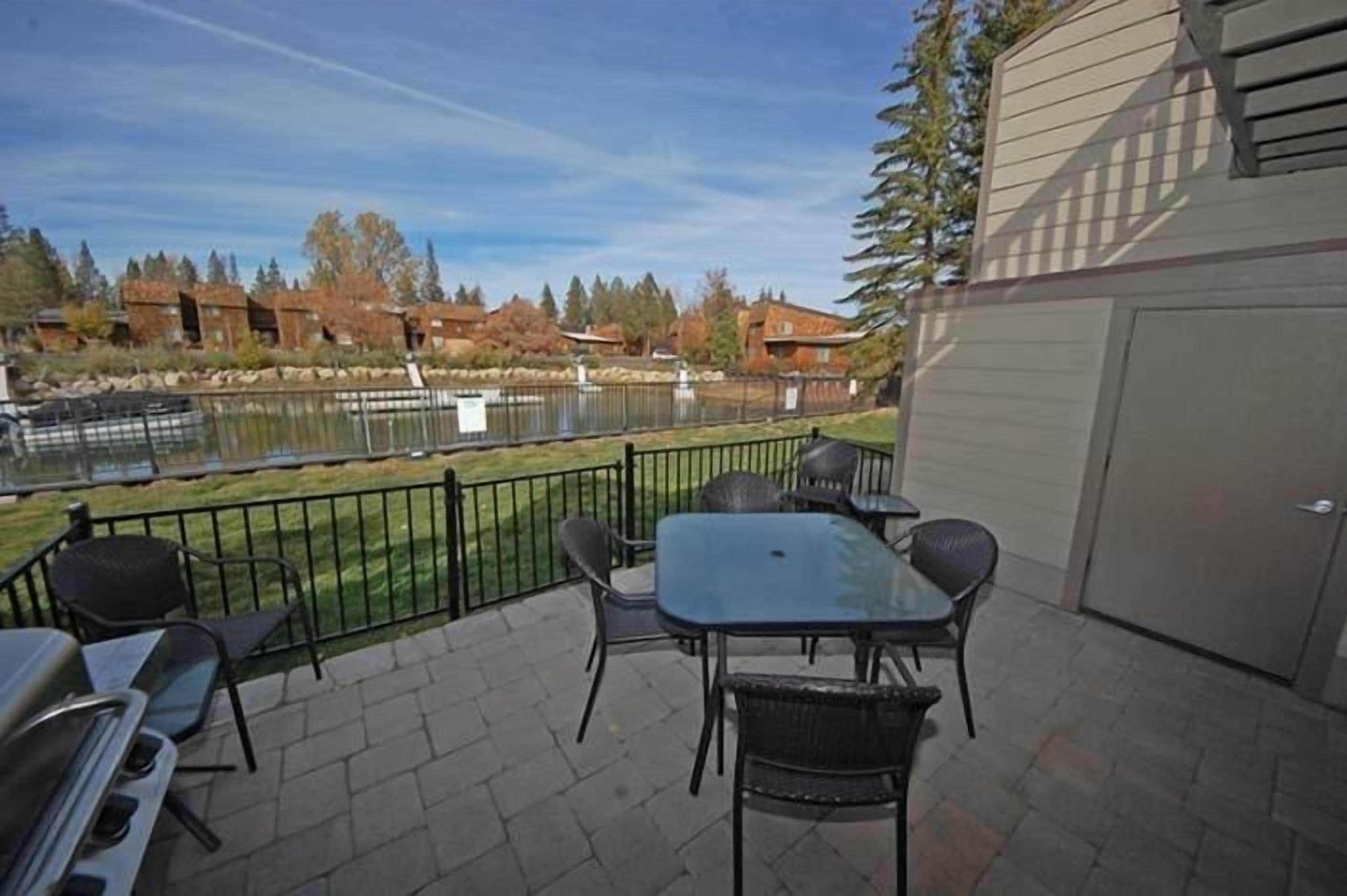 Une terrasse extérieure avec une table et des chaises, un barbecue, et une vue sur un lac et des bâtiments en arrière-plan.