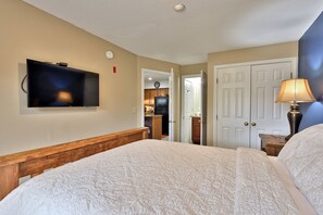 A hotel room with a large bed, a flat-screen TV mounted on the wall, a lamp on a bedside table, and a door leading to a kitchen area. - Pico Mountain Condo F106 3 Bedroom Home (Killington)