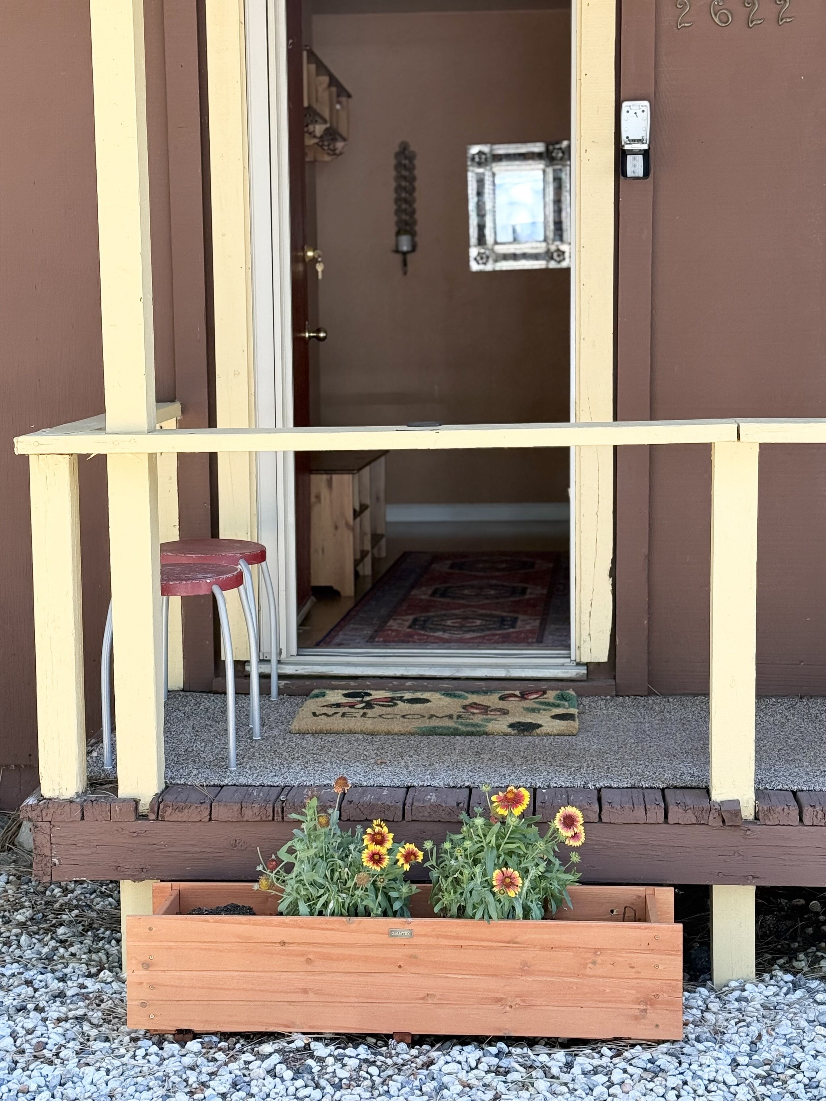 A wooden bench with a red stool, a wooden planter with flowers, and a door leading to an interior space with a rug and a wall-mounted light fixture.