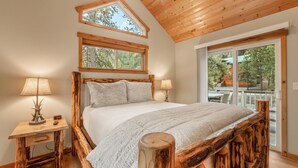 A bedroom with a wooden headboard, a large bed with white bedding, two bedside lamps, and a window overlooking a deck. - Woodlands House 3 Bedroom Home (Truckee)