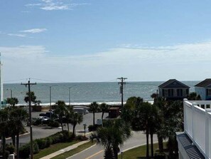 A coastal view with a road, palm trees, and a building with a balcony overlooking the ocean. - Sea Pearl 7 Bedroom Home (Surfside Beach)