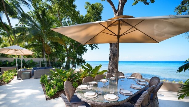 A dining area with a table set for four, surrounded by wicker chairs, under a large umbrella, with a view of the ocean. - Sea Grove House 5 Bedroom Villa (Weston)