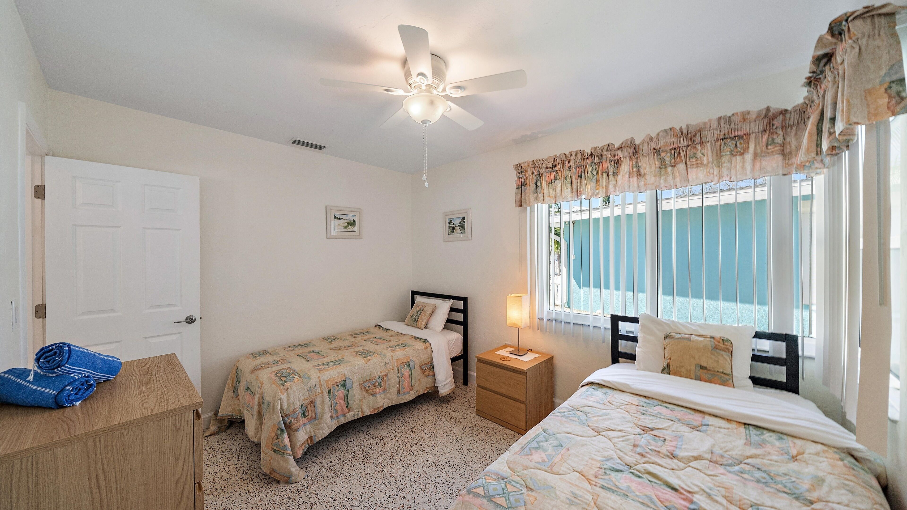 A bedroom with two beds, a ceiling fan, a window with curtains, and a door.
