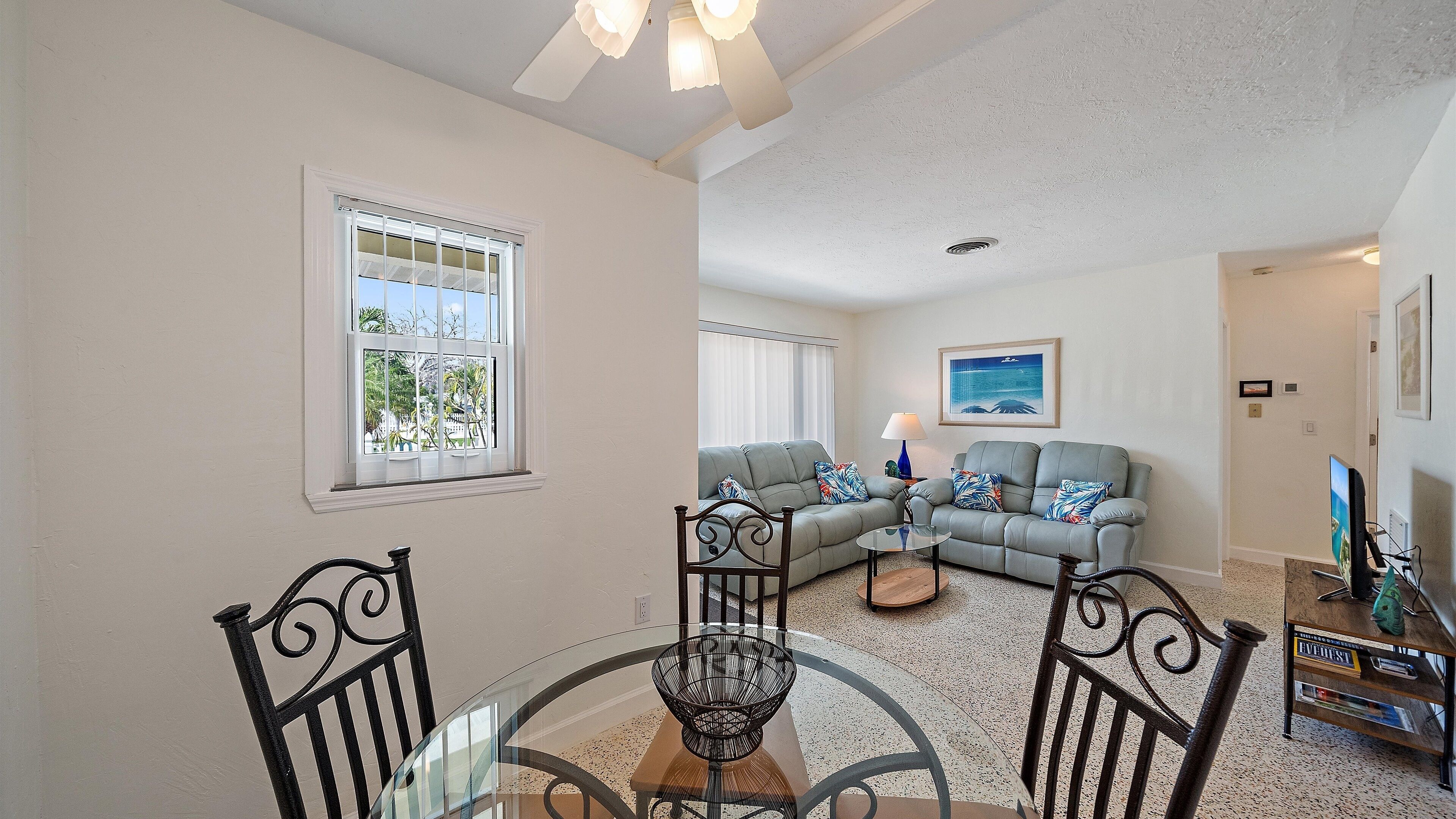 A living room with a grey sofa, a round glass table, and a dining area with chairs.