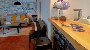 A living room with a piano, a sofa with cushions, a coffee table, and a fireplace with a chalkboard. - House of Rising Sun (Remédios, Ponta Delgada)