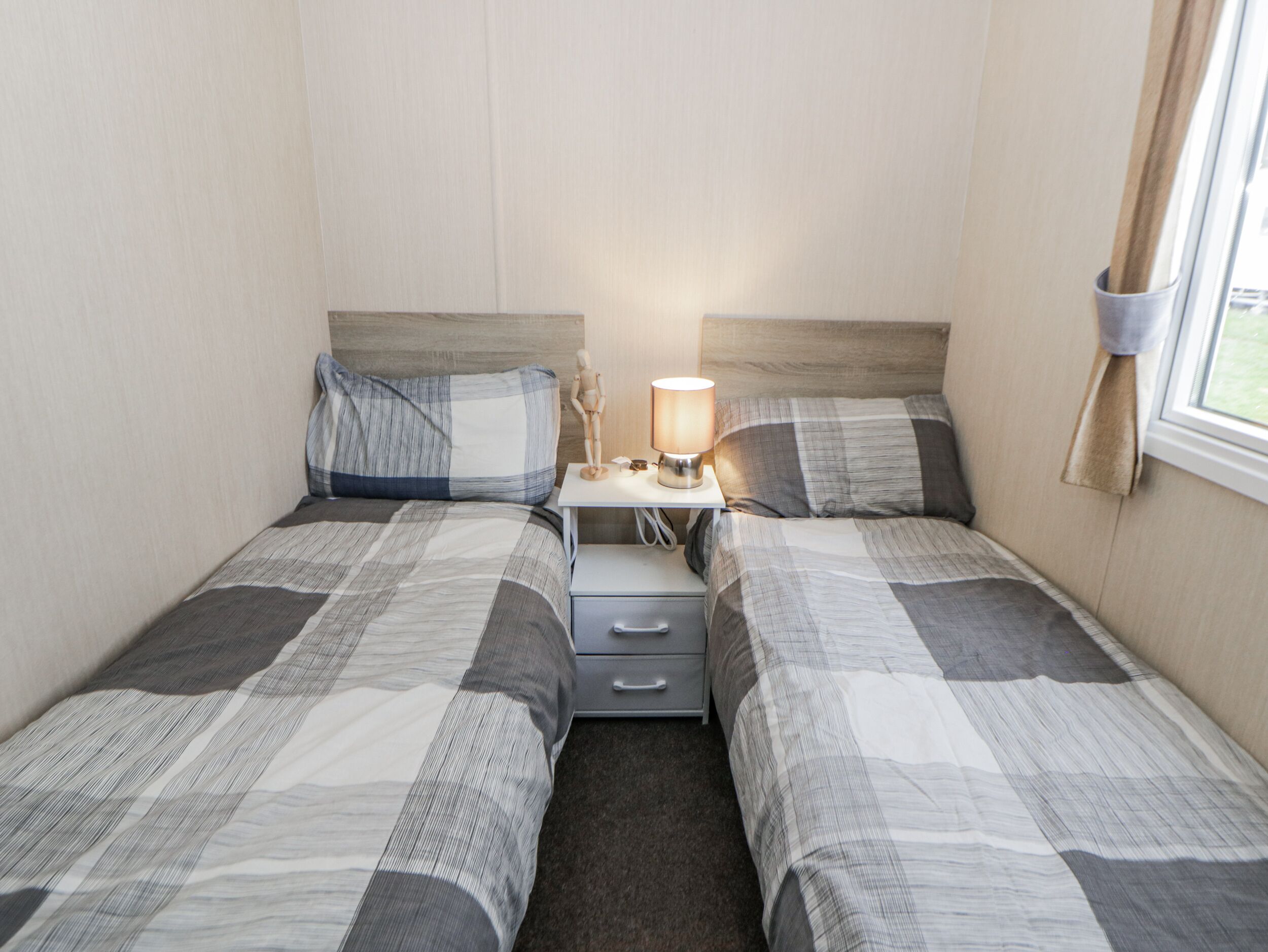 Et tvillingeværelse med to senge, et natbord med lampe og et vindue med gardiner.