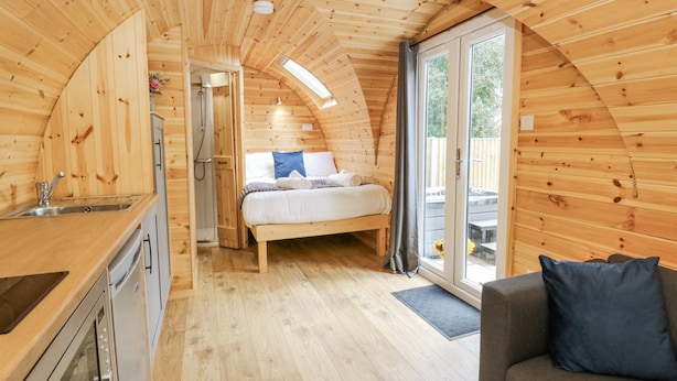 A cozy, wooden cabin interior with a kitchen, a bedroom with a bed and a sofa, and a large sliding glass door leading to an outdoor area. - Oak View (Rhayader)