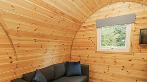 A cozy cabin interior with a curved wooden ceiling, a built-in sofa with cushions, and a window providing a view of greenery outside. - Oak View (Rhayader)