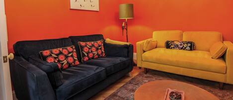 A living room with two sofas, one black and one yellow, against orange walls. There's a patterned rug on the floor and a small round table with a decorative item on it.