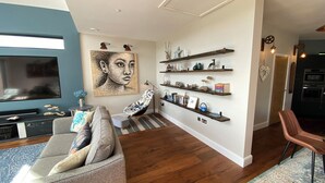 Una sala de estar moderna con un televisor de pantalla plana, un sofá gris, un sillón estampado y un cuadro abstracto grande. Se observan estantes de madera con objetos decorativos y libros.