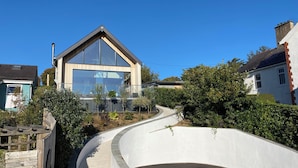 Una casa moderna con un amplio frente de vidrio, una terraza de madera y un camino de acceso curvo.