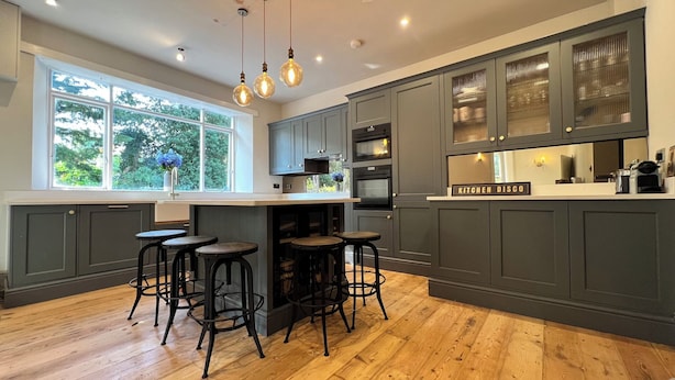 A kitchen with dark grey cabinets, a wooden island with stools, and a window with a view of greenery. - Your Country House (Reading)