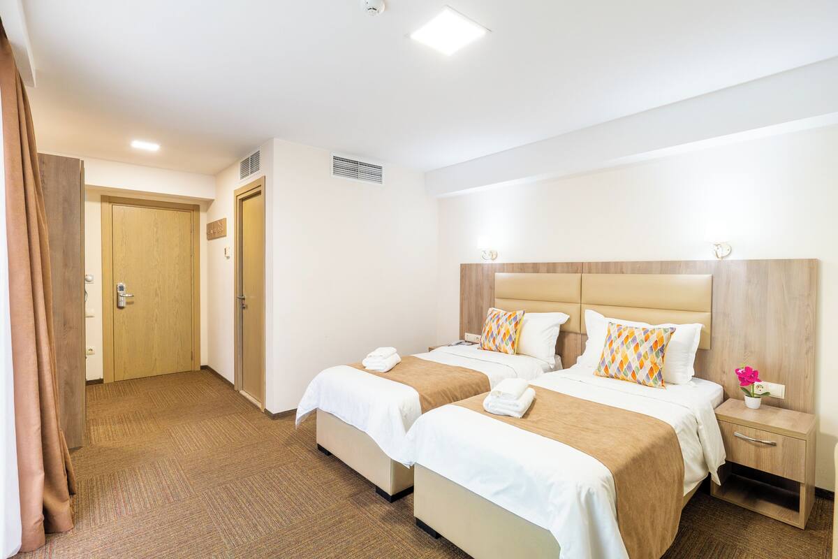 Habitación de hotel con dos camas, cabecera de madera, mesita de noche con lámpara y puerta de baño.