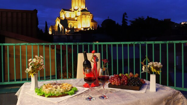 A table set with food and drinks, a bottle of wine, and a vase of flowers, with a building in the background. - Hotel Somi Inn (Tbilisi)