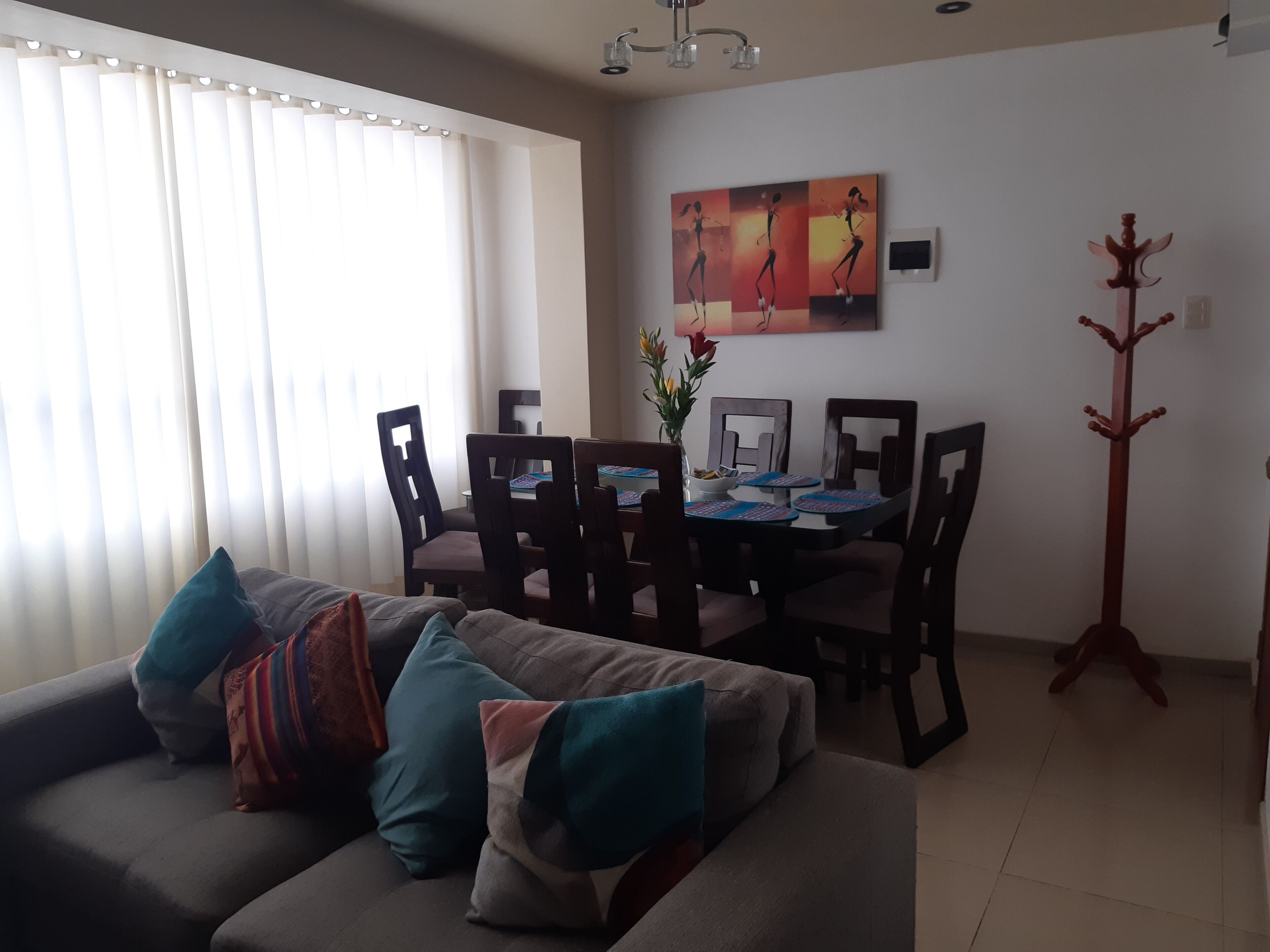 Una sala de estar con un sofá gris, una mesa de comedor con sillas, un cuadro en la pared y un perchero.