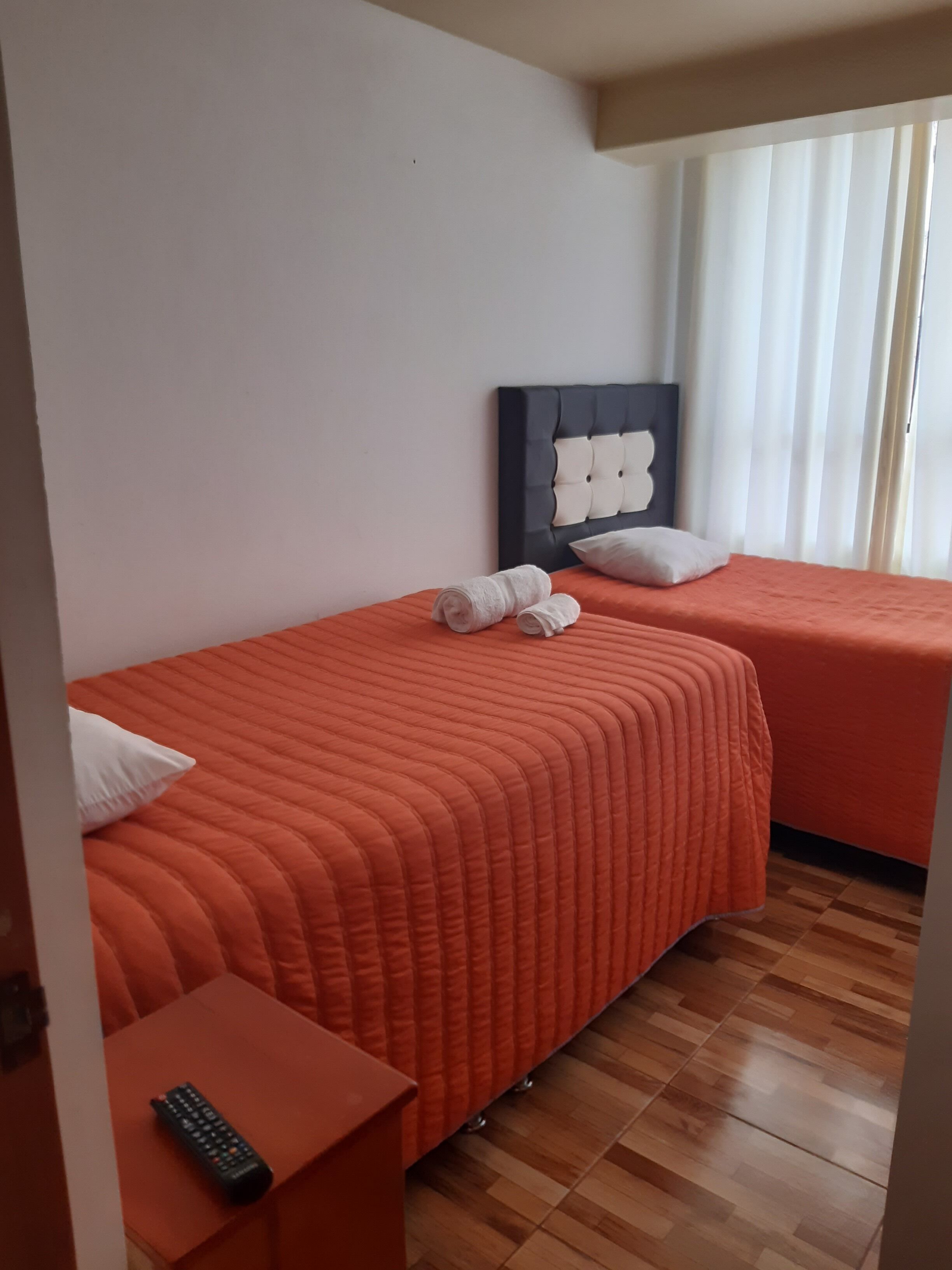 Habitación de hotel con dos camas, cada una con colcha naranja y almohadas blancas. Se observa un cabecero negro con diseño decorativo, una mesita con un control remoto y una ventana con cortinas blancas.