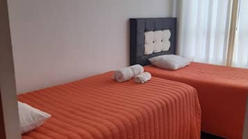Habitación de hotel con dos camas, cada una con colcha naranja y almohadas blancas. Se observa un cabecero negro con diseño decorativo, una mesita con un control remoto y una ventana con cortinas blancas.