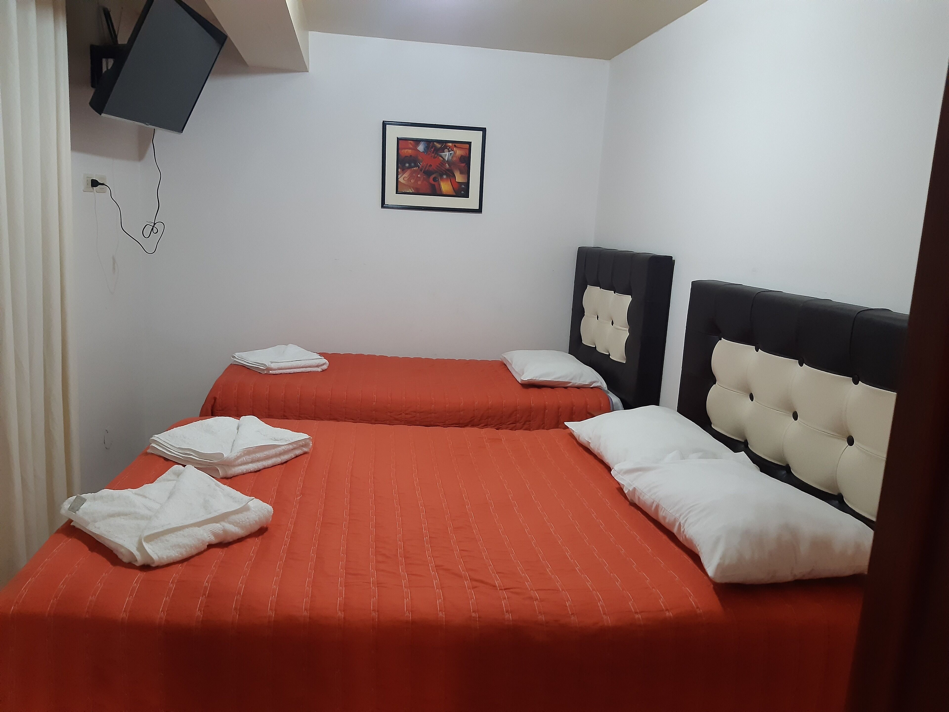 A hotel room with two beds, each with a red bedspread and white pillows. There is a flat-screen TV mounted on the wall and a framed picture above the beds.