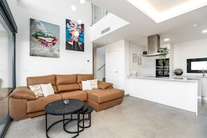 Ein modernes Wohnzimmer mit einem braunen Ecksofa, einem runden schwarzen Tisch und einer Küchenzeile mit weißer Arbeitsplatte und Edelstahlgeräten.