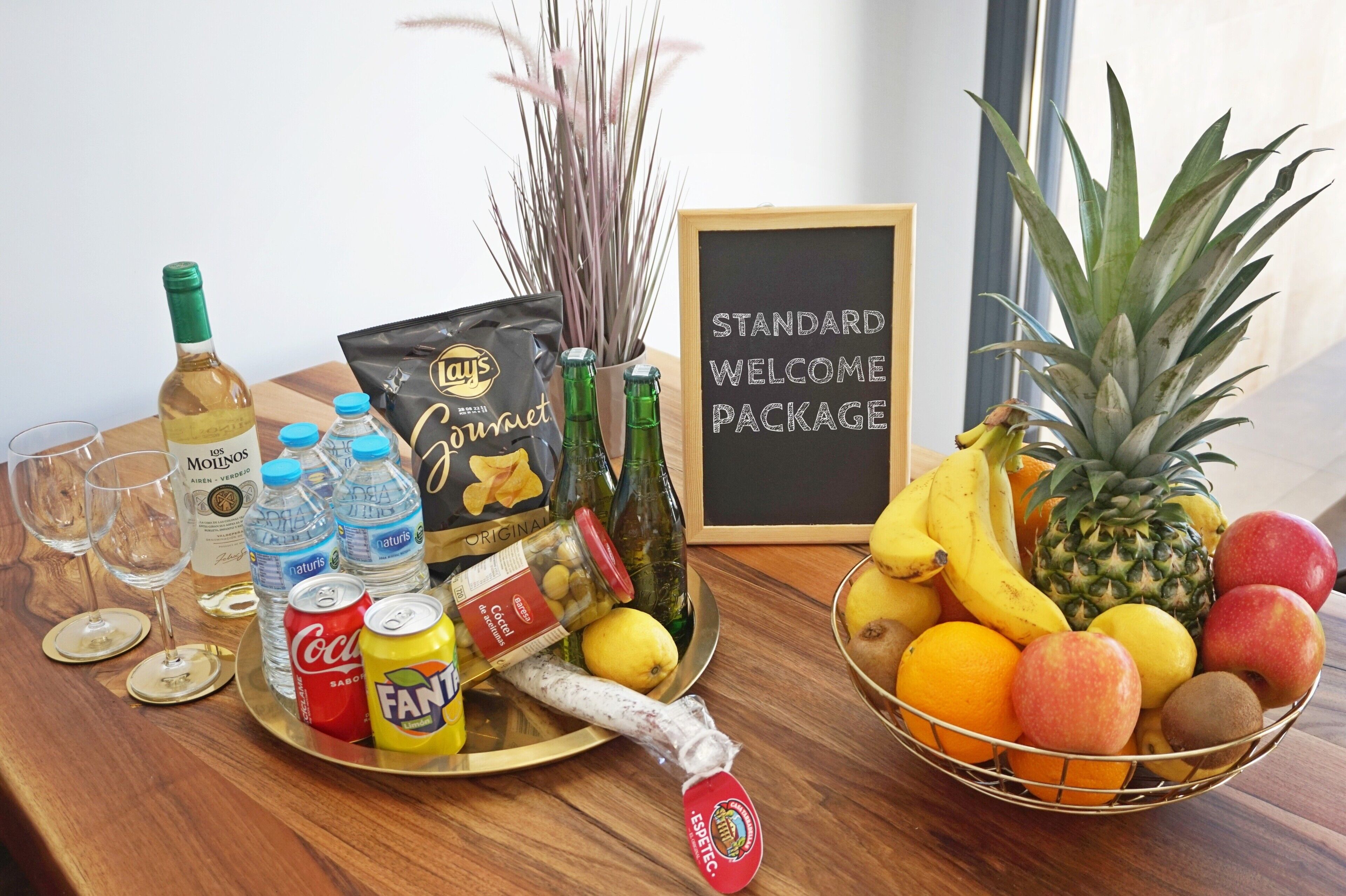 A table with a bottle of wine, water bottles, a can of soda, a bag of snacks, a jar of olives, a bottle of sauce, a bottle of champagne, a bottle of beer, a glass of wine, a tray with a banana, a pineapple, and a bowl of fruit.