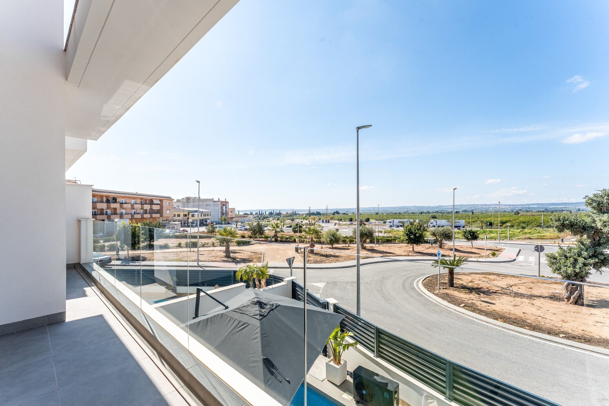A modern balcony with a glass railing, a pool, and a view of a road and distant buildings.