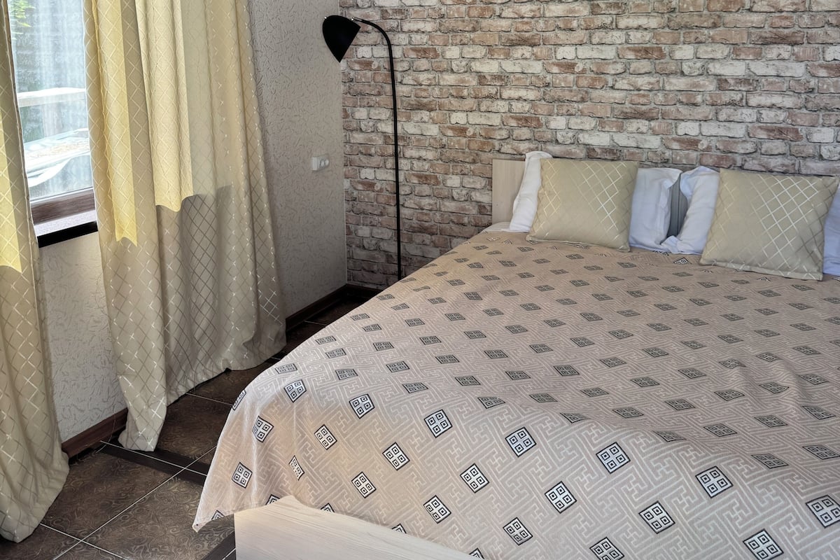 Un dormitorio con pared de ladrillo visto, una cama con ropa de cama estampada, una lámpara de pie y una ventana con cortinas.