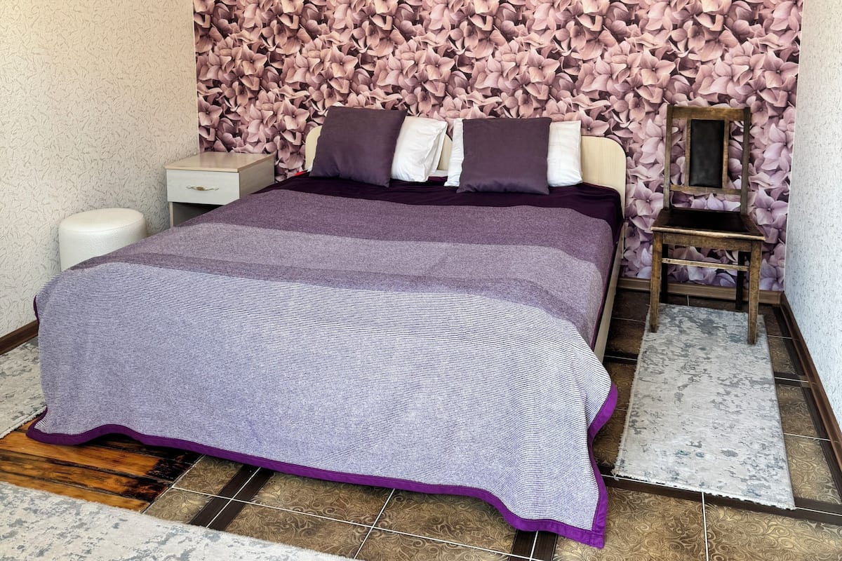 Un dormitorio con cama, mesitas de noche, una silla, un estante en la pared y un papel tapiz estampado.