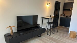Moderni olohuone, jossa on litteänäyttöinen televisio, pieni pöytä, kaksi baarituolia ja koristeellinen palmu.