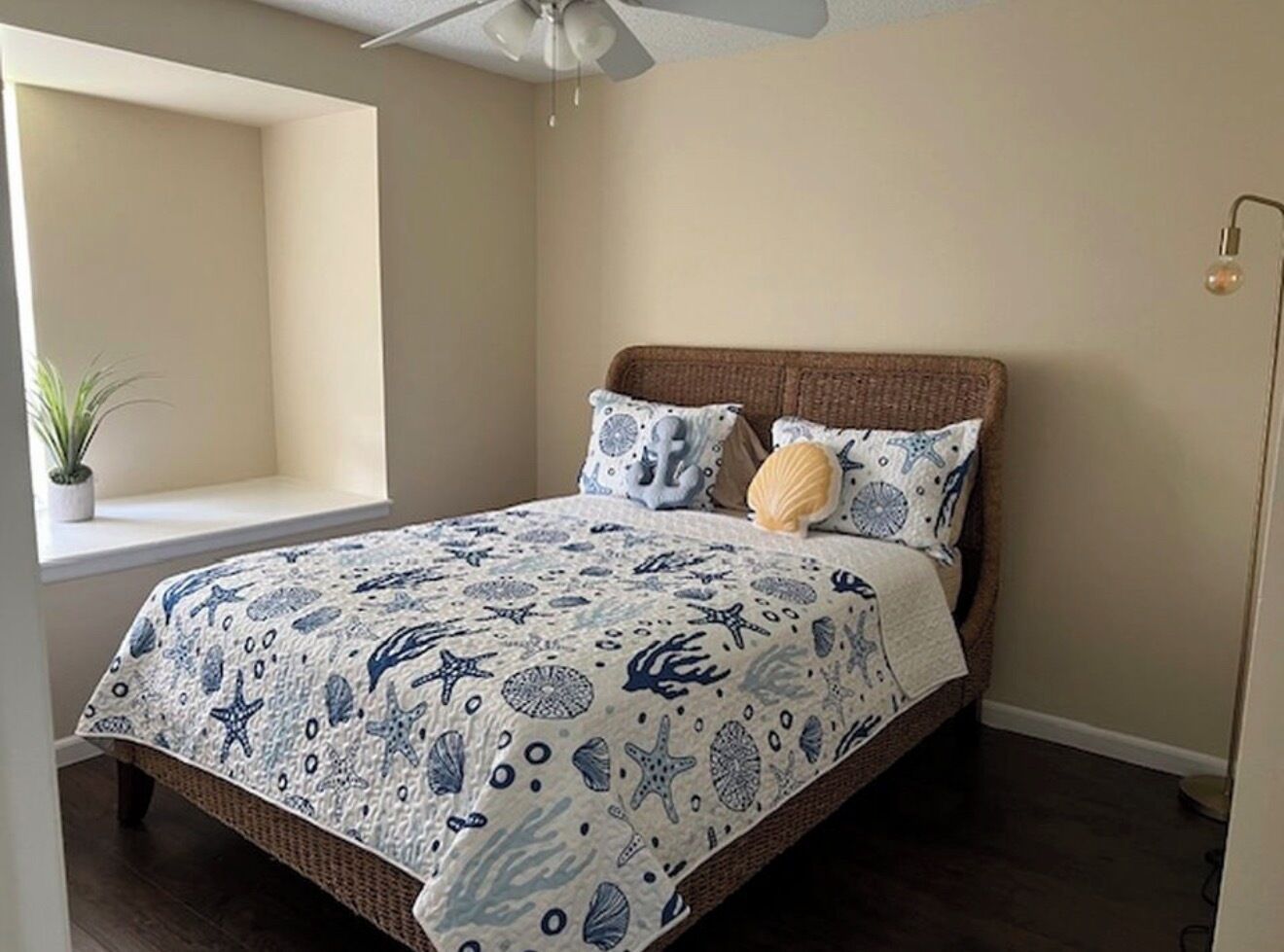 Une chambre à coucher avec un lit, un ventilateur de plafond, une fenêtre avec une plante et un luminaire fixé au mur.