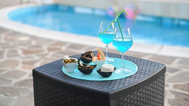 A poolside table with a wicker chair, a glass of blue cocktail, and a selection of seafood and olives. - Villa with a pool, Puglia – Libellule 3 (Carovigno)