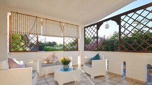A sunlit patio with white wicker furniture, a small table with a vase of flowers, and lattice window frames. - Villa with a pool, Puglia – Libellule 3 (Carovigno)