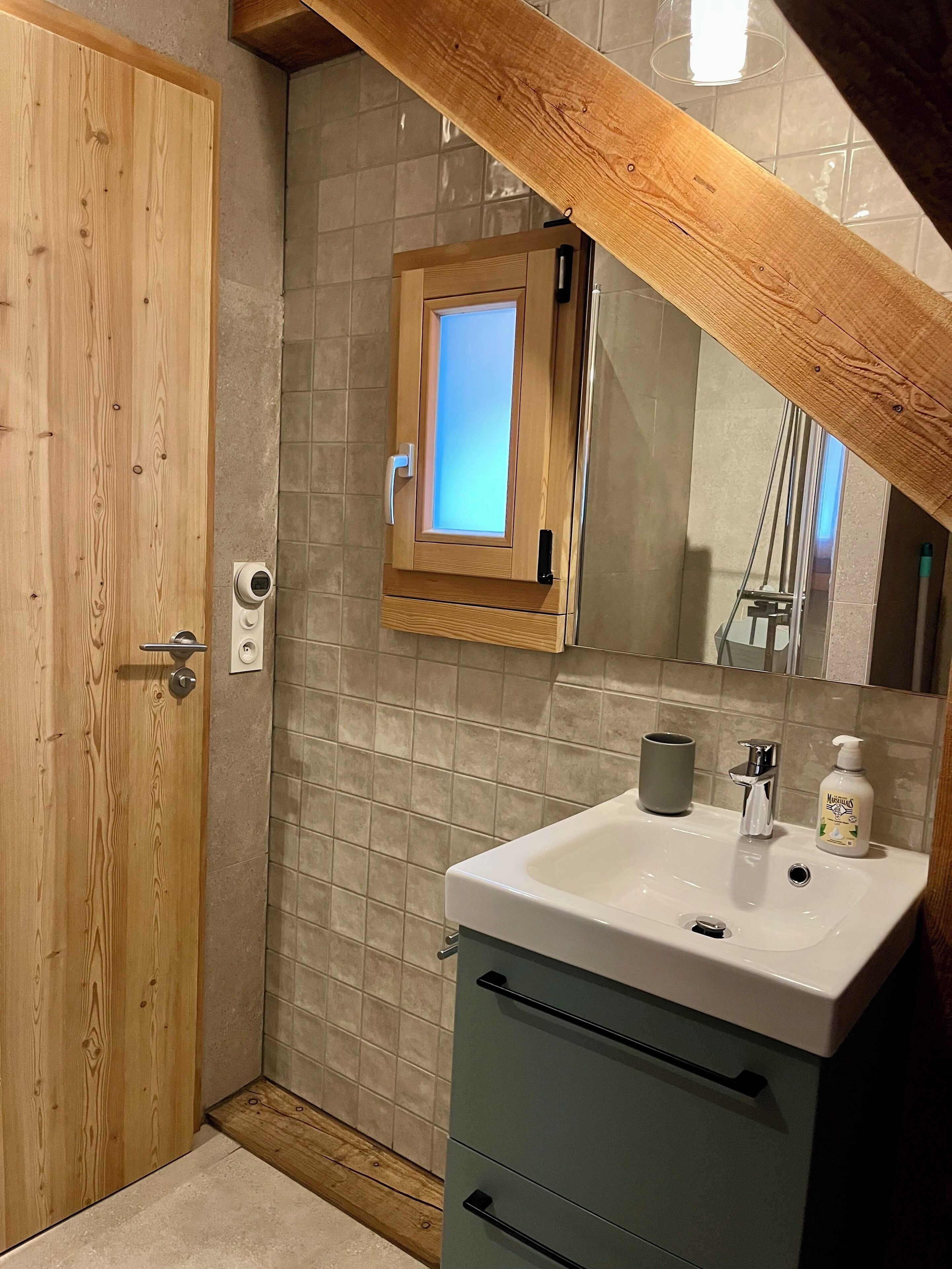 A bathroom with a wooden door, a tiled wall, a small window, a sink with a mirror, and a green cabinet.