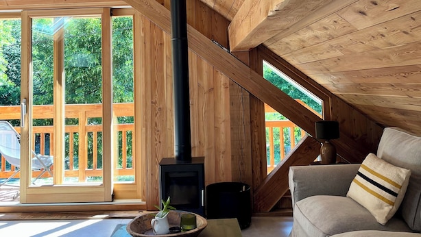 Ein gemütliches Schlafzimmer im Dachgeschoss mit Holzbalken, einem schwarzen Holzofen, einer Couch und Blick auf eine Holzterrasse.