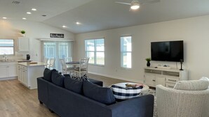 Ein modernes Wohnzimmer mit einem dunkelblauen Sofa, einem weißen Sessel, einem Essbereich mit weißen Stühlen, einer Küche mit weißen Schränken und einem Deckenventilator.