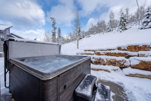 Ein Whirlpool in verschneiter Winterlandschaft mit Steinmauer und Bäumen im Hintergrund.