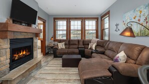 A modern living room with a stone fireplace, leather sectional sofa, and large windows offering a view of the outdoors. - Kicking Horse Buckskin 3-205 Family friendly 2 bed private deck & mountain views (Granby)