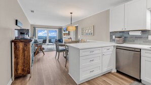 A modern kitchen with white cabinetry, a built-in dishwasher, and a dining area with a view. - Newly Renovated Oceanfront 3BR at Sea Cloister ll | Cherry Grove (North Myrtle Beach)