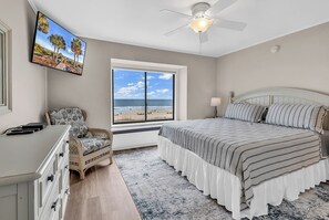 A bedroom with a bed, a desk, a chair, a TV, a ceiling fan, a window with a beach view, and a rug. - Newly Renovated Oceanfront 3BR | Top Floor (North Myrtle Beach)