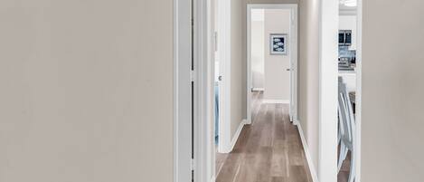 A hallway with a wooden floor, white walls, and a door at the end leading to a kitchen area.