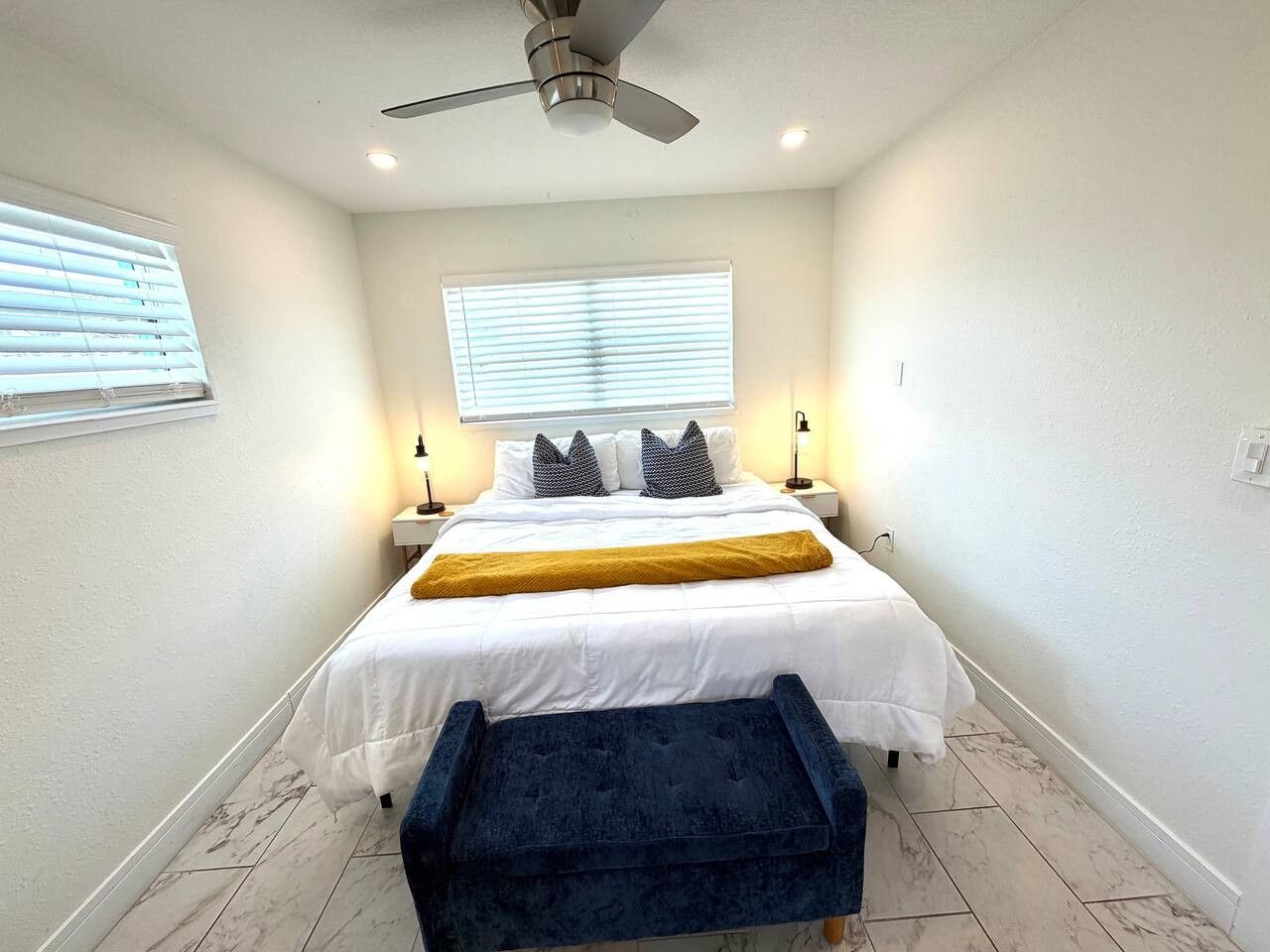 Un dormitorio con cama, lámparas de noche, ventilador de techo y ventana con persianas.
