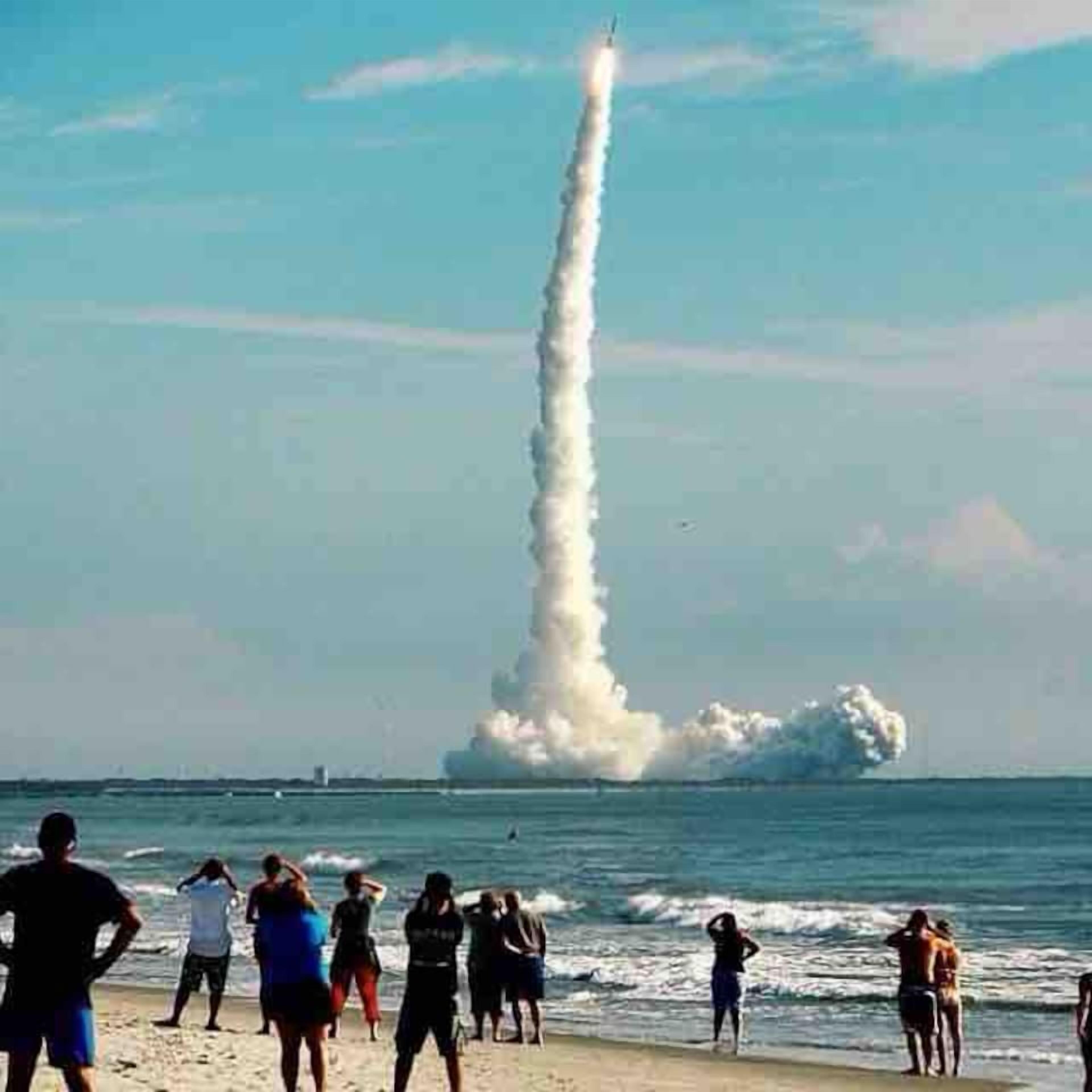 Escena de playa con un lanzamiento de cohete, espectadores en la orilla y el océano al fondo.