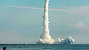 Escena de playa con un lanzamiento de cohete, espectadores en la orilla y el océano al fondo.